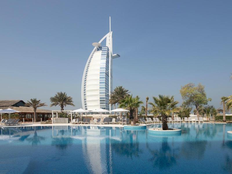 Moderner Wolkenkratzer mit Segelform vor blauem Himmel, umgeben von Palmen und großem Schwimmbecken.