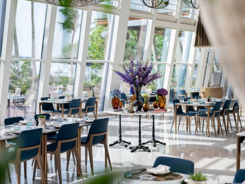 Helles Café mit großem Glasfenster, blauen Stühlen, Holztischen und dekorativen Blumen auf Tischen.