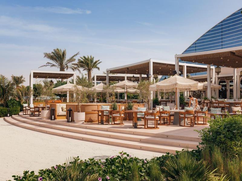 Moderner Außenbereich eines Restaurants mit Holzmöbeln, Sonnenschirmen und Palmen bei blauem Himmel.