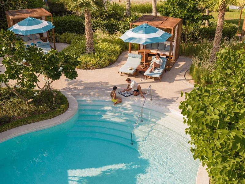 Pool mit klarstem Wasser, Palmen, Liegestühlen und blauen Sonnenschirmen in tropischer Umgebung.