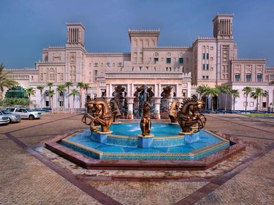 Großes Sandsteingebäude mit drei Türmen und bronzenen Statuen in blauem Brunnen davor.