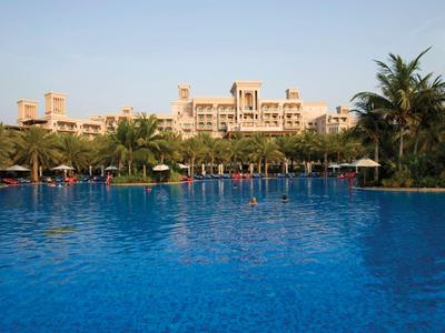 Großer blauer Swimmingpool vor einem Hotelgebäude mit Palmen und Sonnenschirmen.