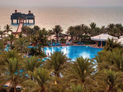 Blick auf Hotelpool umgeben von Palmen und Strand mit Steg und blauem Meer im Abendlicht.