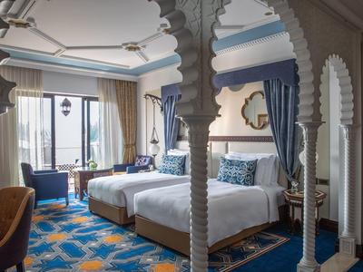 Hell eingerichtetes Zimmer mit zwei Betten, blauer Teppich, und großen Fenstern mit blauen Vorhängen.