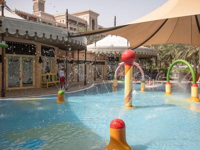 Kinderspielplatz mit Wasserfontänen, bunten Figuren und einem schattigen Zelt über einem kleinen Pool.