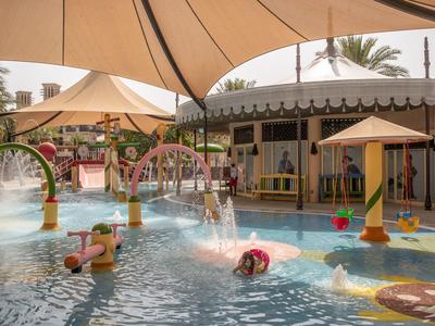 Kindergarten-Wasserspielplatz mit bunten Rutschen, Schirmen und Wasserspielen unter Sonnensegeln.