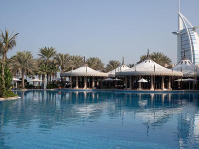 Blauer Pool mit Palmen und weißen Sonnenschirmen, Burj Al Arab Hotel im Hintergrund bei klarem Himmel.
