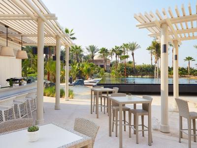 Hell gestaltete Außenbar mit weißen Möbeln, Holzstruktur und Palmen vor blauem Himmel und Wasserfläche.