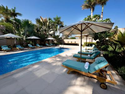 Poolbereich mit blauer Wasserfläche, Liegestühlen mit blauen Polstern und großen Sonnenschirmen.