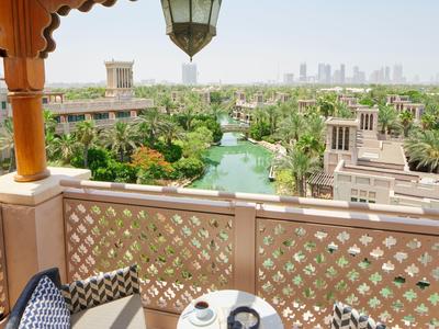 Balkon mit weißen Stühlen und kleinem Tisch, grüne Gartenanlage und Stadt im Hintergrund.