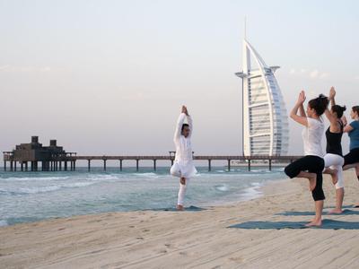 Vier Personen praktizieren Yoga am Strand mit Hafen und modernem Segelgebäude im Hintergrund.