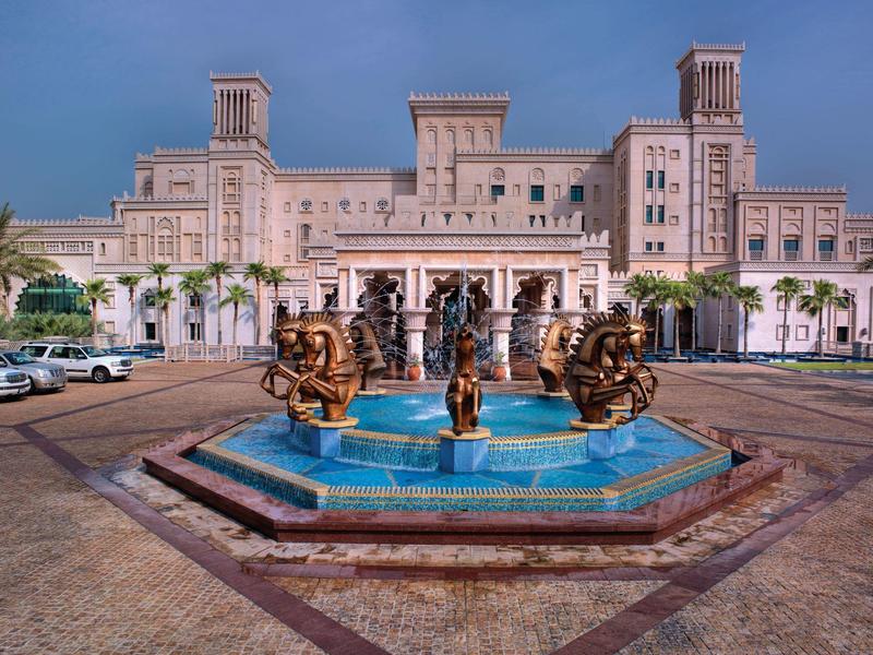 Großes Sandsteingebäude mit drei Türmen und bronzenen Statuen in blauem Brunnen davor.