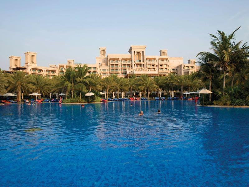 Großer blauer Pool mit Palmen und beige Hotelgebäude im Hintergrund unter klarem Himmel.