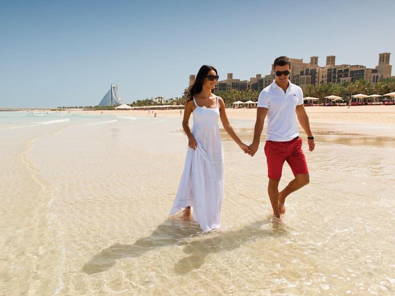 Paar in Weiß dressiert barfuß am flachen Strand mit klarem Wasser und Gebäuden im Hintergrund.