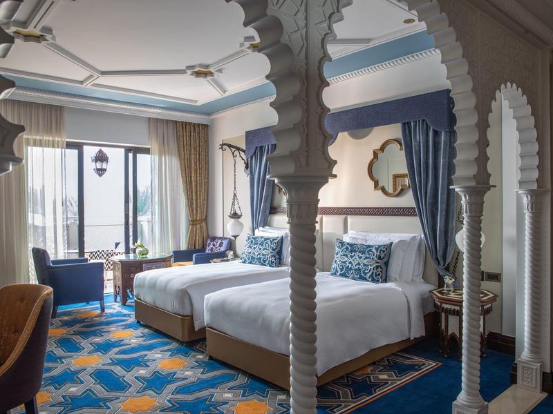 Hell eingerichtetes Zimmer mit zwei Betten, blauer Teppich, und großen Fenstern mit blauen Vorhängen.