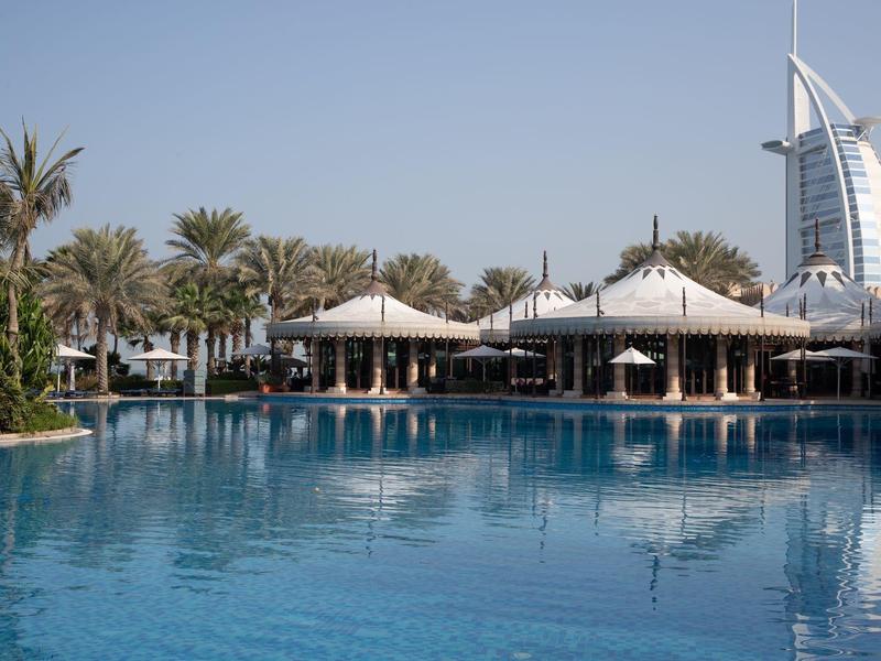 Blauer Pool mit Palmen und weißen Sonnenschirmen, Burj Al Arab Hotel im Hintergrund bei klarem Himmel.