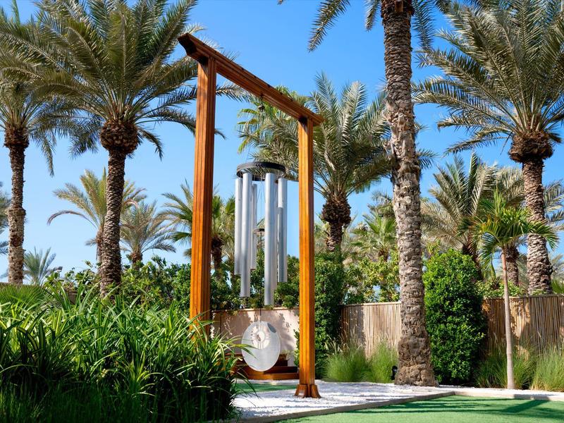 Große Windspiele aus Metall hängen an einem Holzrahmen, umgeben von Palmen und grünen Pflanzen.