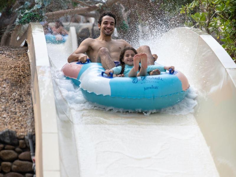 Familie fährt lachend auf blauer Reifenrutsche im Wasserpark, umgeben von Bäumen und Felsen.