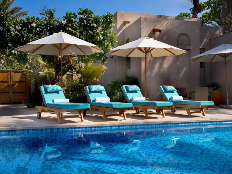 Vier türkisfarbene Liegestühle mit weißen Sonnenschirmen am blauen Pool vor hellen Gebäuden.