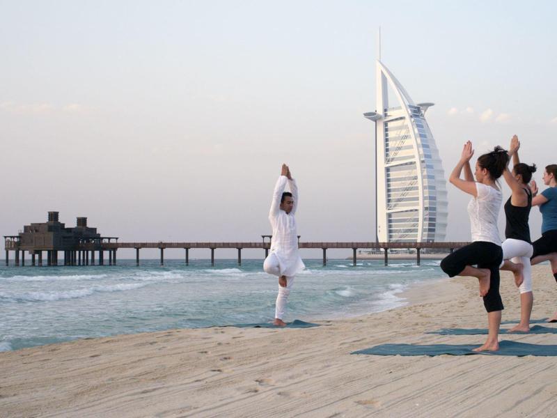 Vier Personen praktizieren Yoga am Strand mit Hafen und modernem Segelgebäude im Hintergrund.