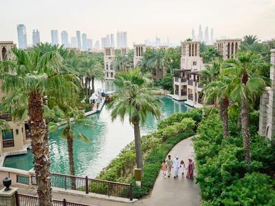 Grüner Kanal mit Palmen, Fußweg und traditionellen Gebäuden vor moderner Stadt mit Hochhäusern.