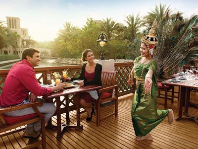 Zwei Personen sitzen an einem Holztisch im Freien, während eine Tänzerin in grünem Kleid mit Pfauenfedern performt.