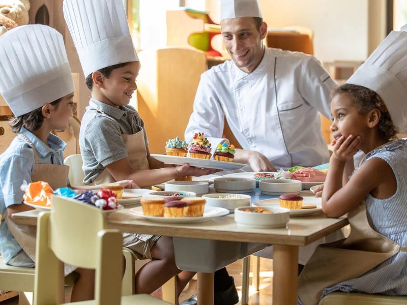 Kinder mit Kochmützen beim Backen, ein Koch lächelt und erklärt, bunte Muffins auf dem Tisch.
