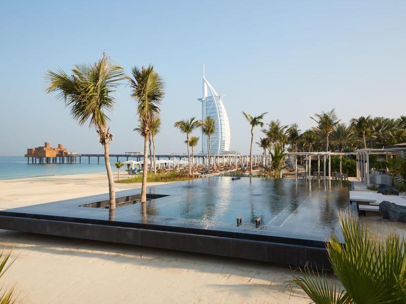 Schwimmbad am Strand mit Palmen, Sonnenschirm und dem Burj Al Arab Hotel im Hintergrund bei klarem Himmel.