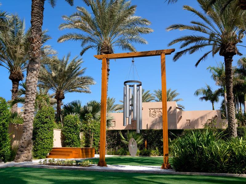 Grüner Garten mit Palmen, Holzbalken und Metall-Glockenspiel vor einem hellen Gebäude.