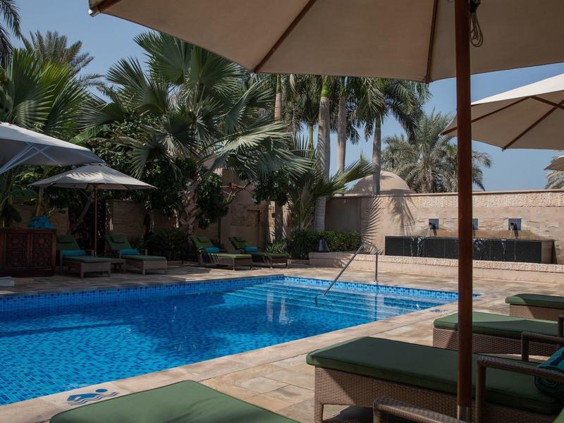 Schwimmbad mit blauen Wasser, Liegestühlen und Sonnenschirmen, umgeben von Palmen und Steinmauern.