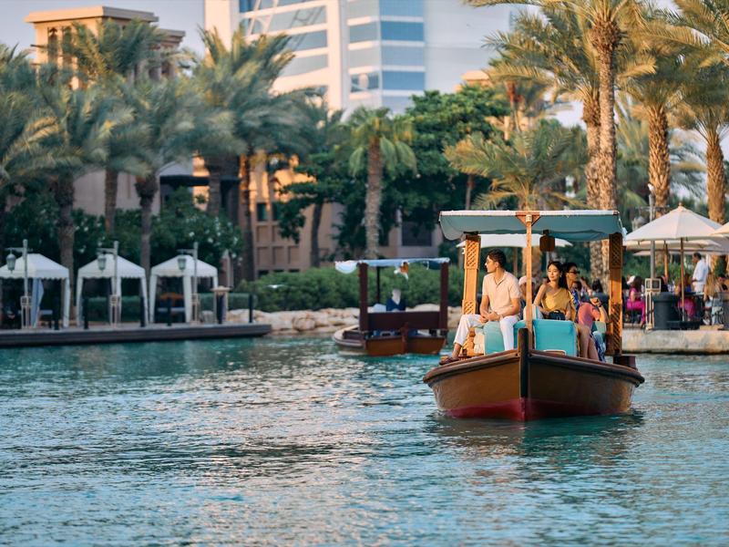 Boot mit Menschen auf Wasser vor Palmen und Gebäuden bei sonnigem Wetter in der Stadt.