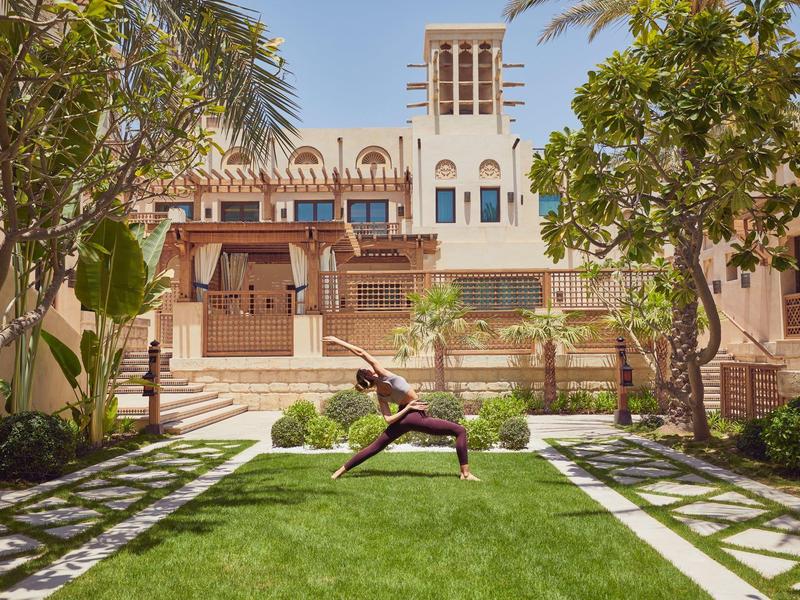 Person macht Yoga-Pose auf grünem Rasen, umgeben von Bäumen und traditioneller Gebäudearchitektur.