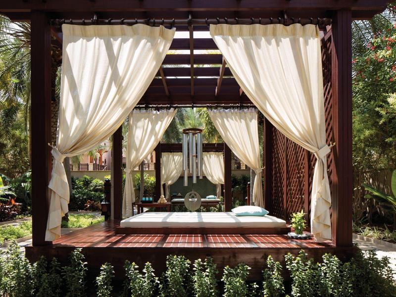 Offener Pavillon mit weißen Vorhängen, Betten und grüner Gartenumgebung bei Sonnenschein.