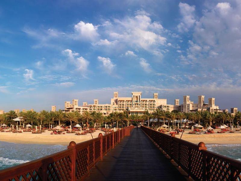 Blick auf einen langen Steg zum Strand mit Palmen und großen Hotelgebäuden unter blauem Himmel.