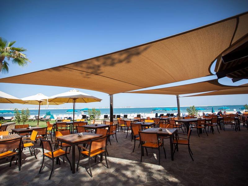 Freiluft-Café mit Holztischen, Stühlen, Sonnensegeln und Blick aufs Meer an einem sonnigen Tag.