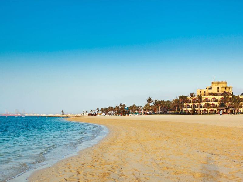 Strand mit goldenem Sand, blauem Meer und Gebäuden im Hintergrund unter klarem Himmel.