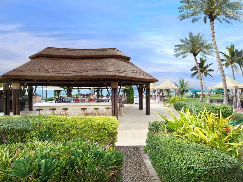 Offene Holzhütte mit Strohdach, umgeben von grünen Hecken und Palmen unter blauem Himmel.