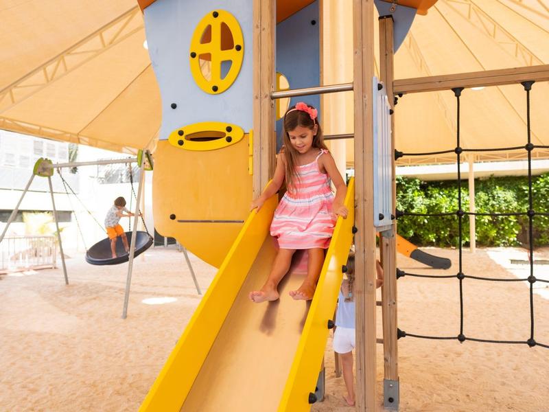 Mädchen rutscht eine gelbe Rutsche auf einem Spielplatz mit Schaukeln und Klettergerüst.