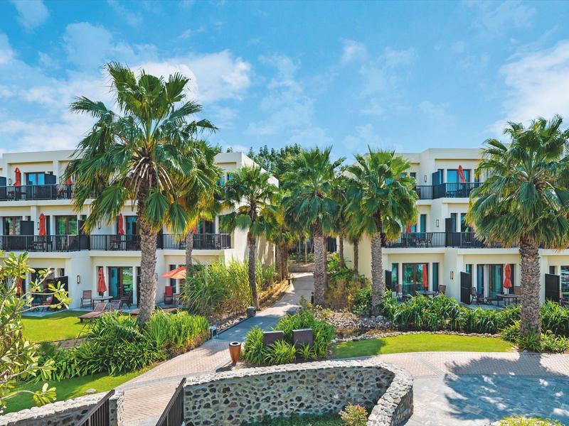 Modernes Hotelgebäude mit Balkonen, Palmen und gepflegtem Garten unter blauem Himmel.