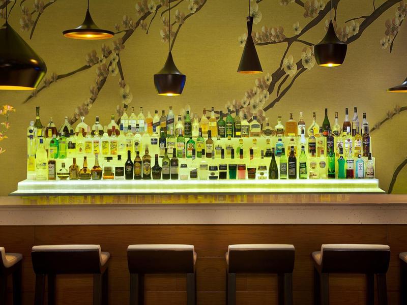 Moderne Bar mit beleuchtetem Regal, vielen Flaschen und fünf Hockern vor dem Tresen.