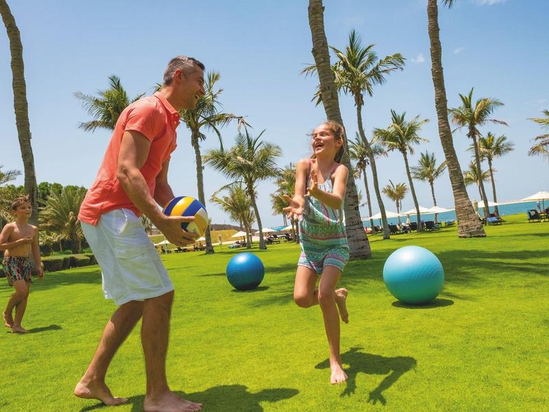 Menschen spielen Volleyball auf grünem Rasen mit Palmen und blauem Himmel im Hintergrund.