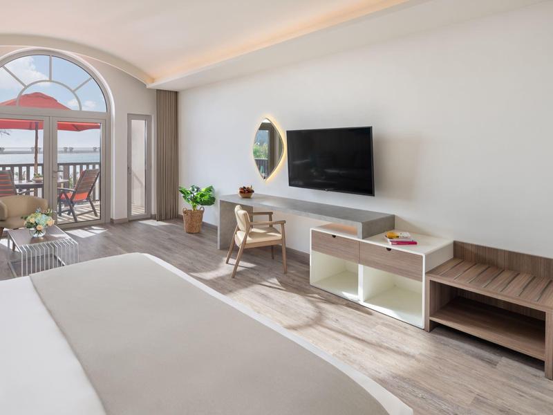 Helles Hotelzimmer mit großem Bett, Fernseher, Schreibtisch und Balkon mit Sitzbereich und rotem Sonnenschirm.