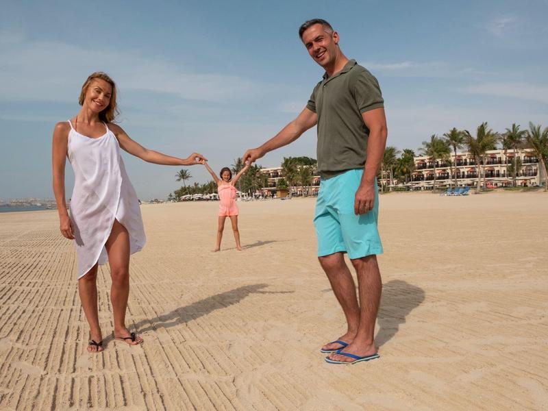 Zwei Erwachsene halten Hände am Strand, ein Kind läuft im Hintergrund, Palmen und Gebäude sichtbar.