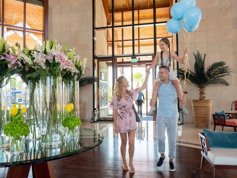 Familie betritt helles Zimmer mit großen Blumenvasen, Holzfußboden und blauen Luftballons.