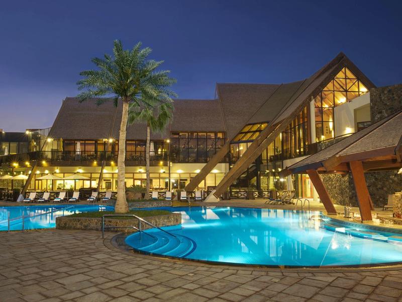 Beleuchteter Poolbereich mit Liegestühlen, Palmen und modernem Hotelgebäude bei Dämmerung.