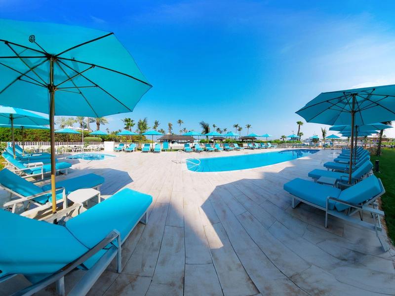 Großer Außenpool mit blauen Liegen und Sonnenschirmen, umgeben von Palmen und strahlend blauem Himmel.