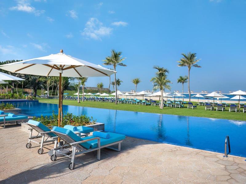 Schwimmbad mit Sonnenliegen, weißen Sonnenschirmen, Palmen und Blick auf das Meer unter blauem Himmel.