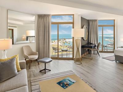 Habitación de hotel luminosa con balcón y vistas al mar, muebles modernos y ambiente acogedor.