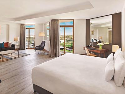 Habitación de hotel espaciosa con cama, área de estar, escritorio y balcón con vista a zonas verdes.