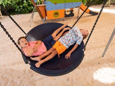 Dos niños descansan en un columpio redondo grande en un área de juegos de arena.